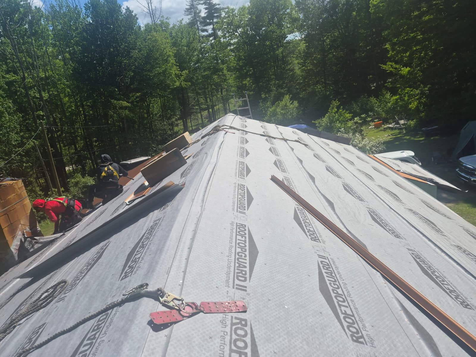 Roof under repair with gray underlayment and workers’ tools, surrounded by trees in bright sunlight
