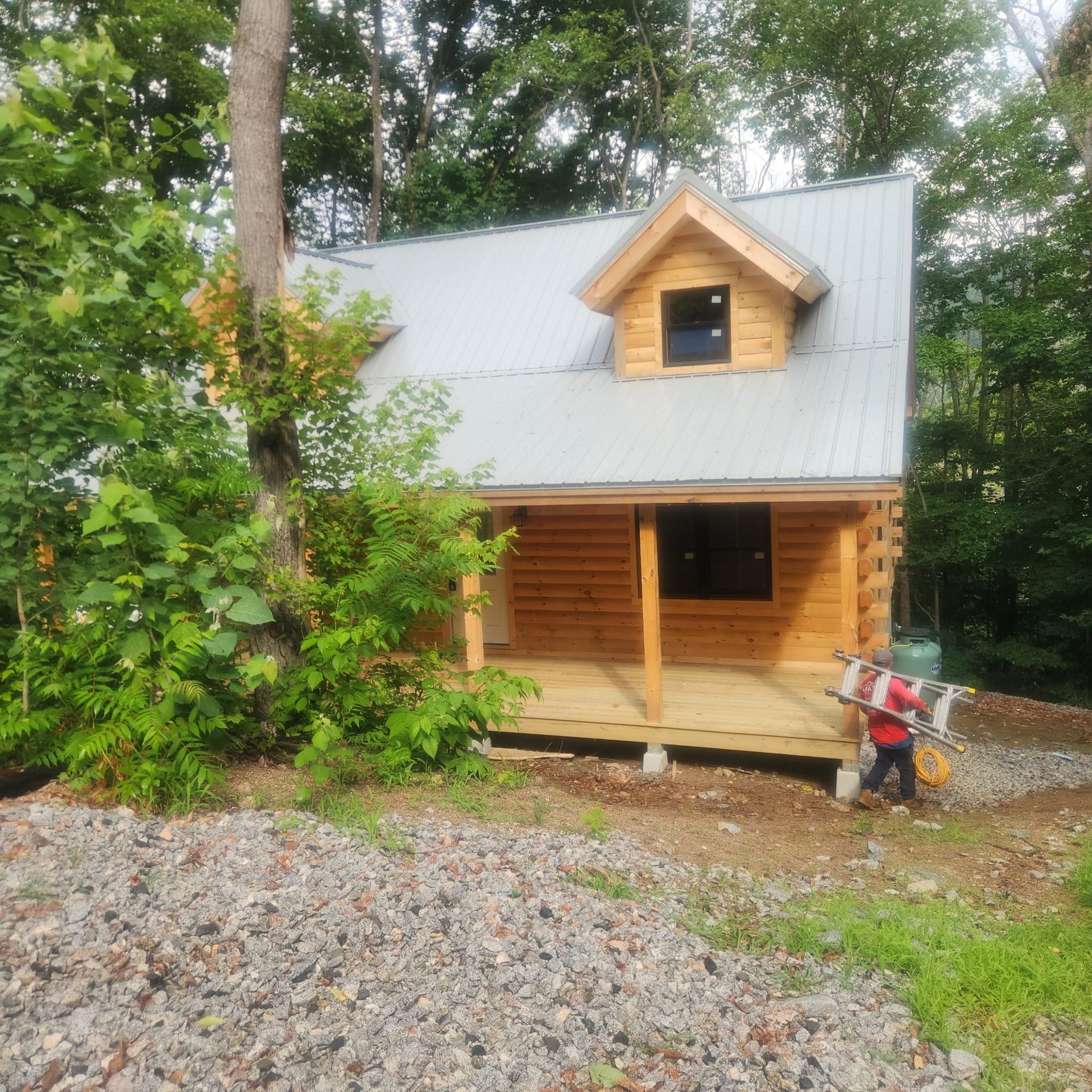 Wooden cabin with metal roof in a forest, with a person carrying a child on the right.