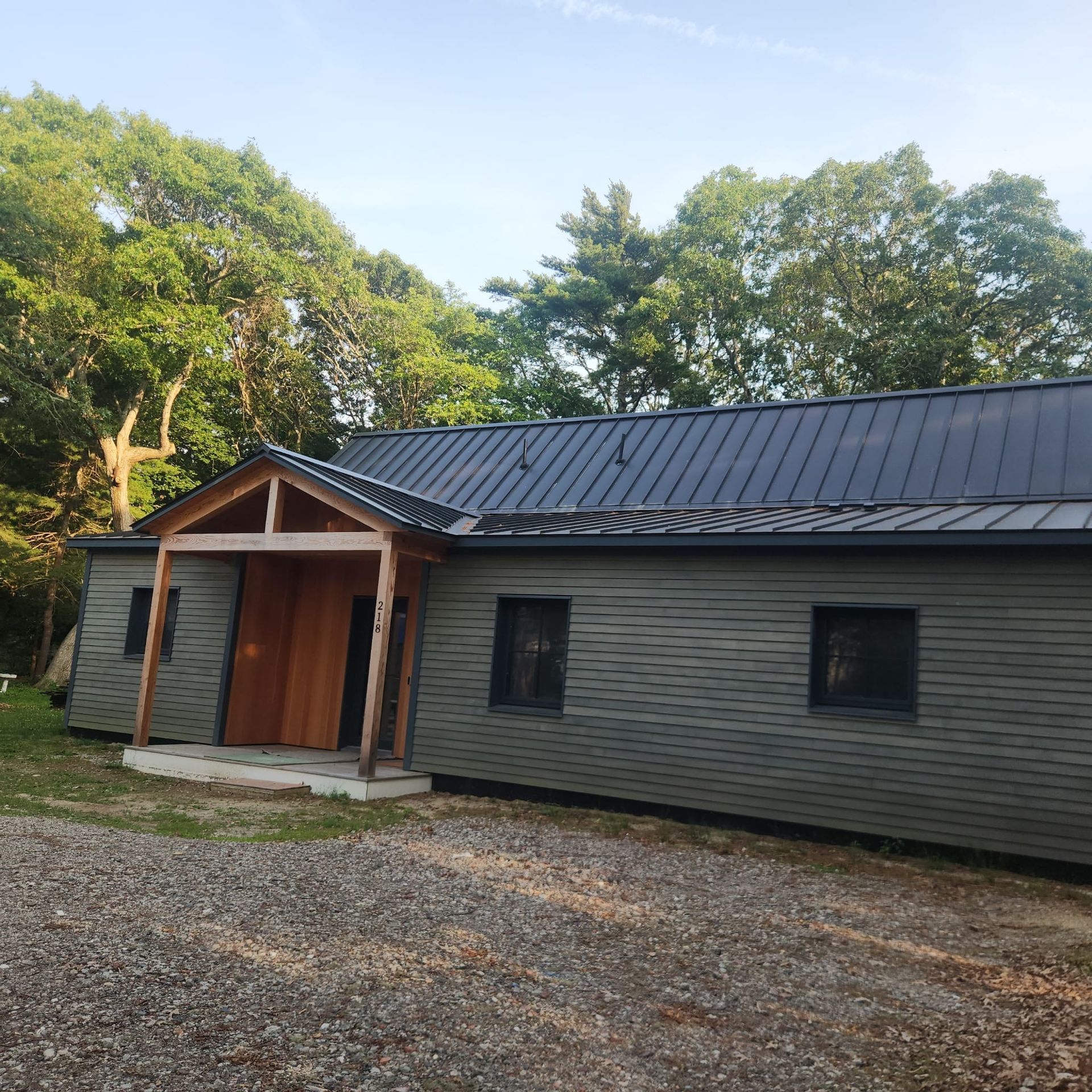 Single-story gray cabin with black roof, wooden entry porch, and trees behind it.