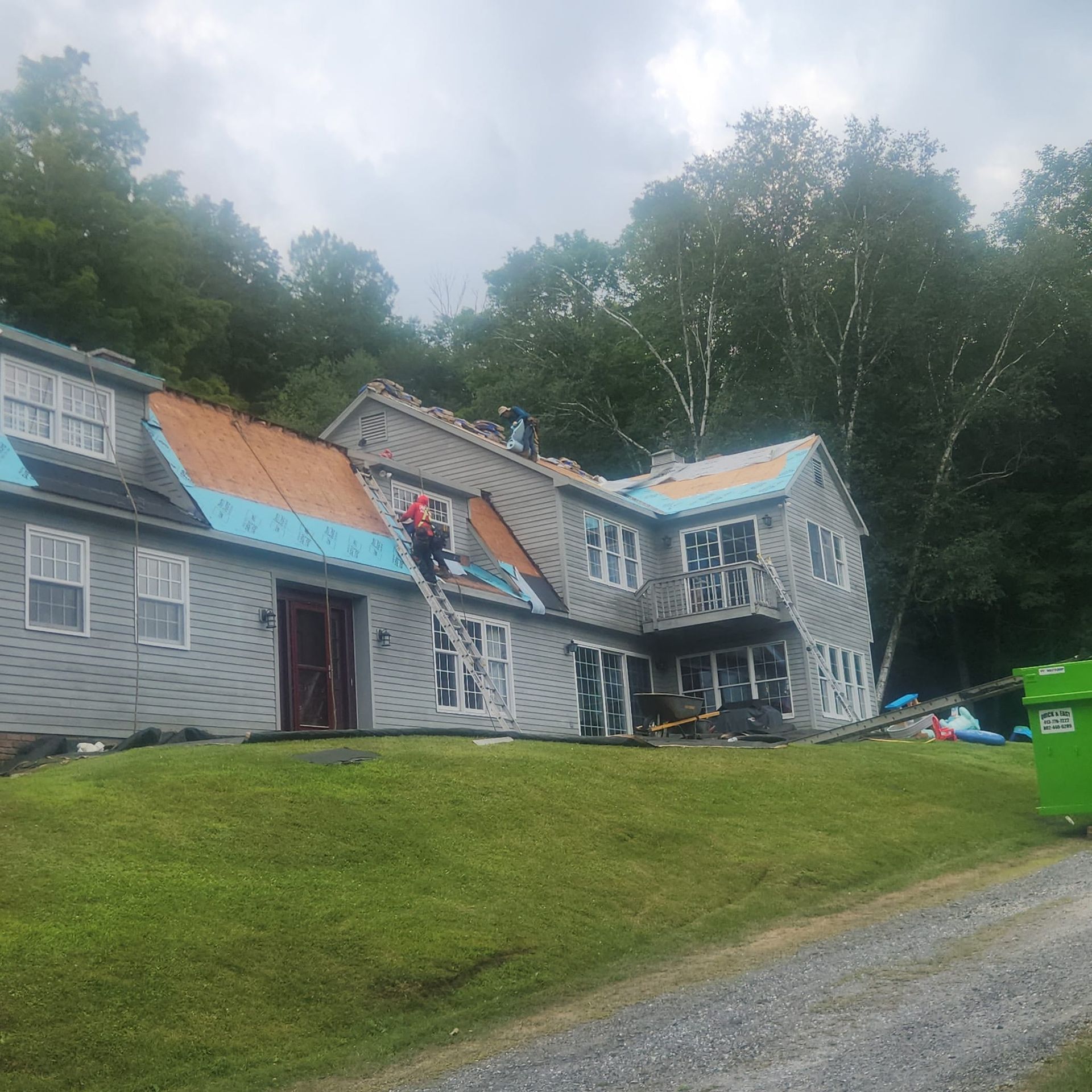 Damaged gray house with a caved-in roof and blue tarps on a grassy slope near trees