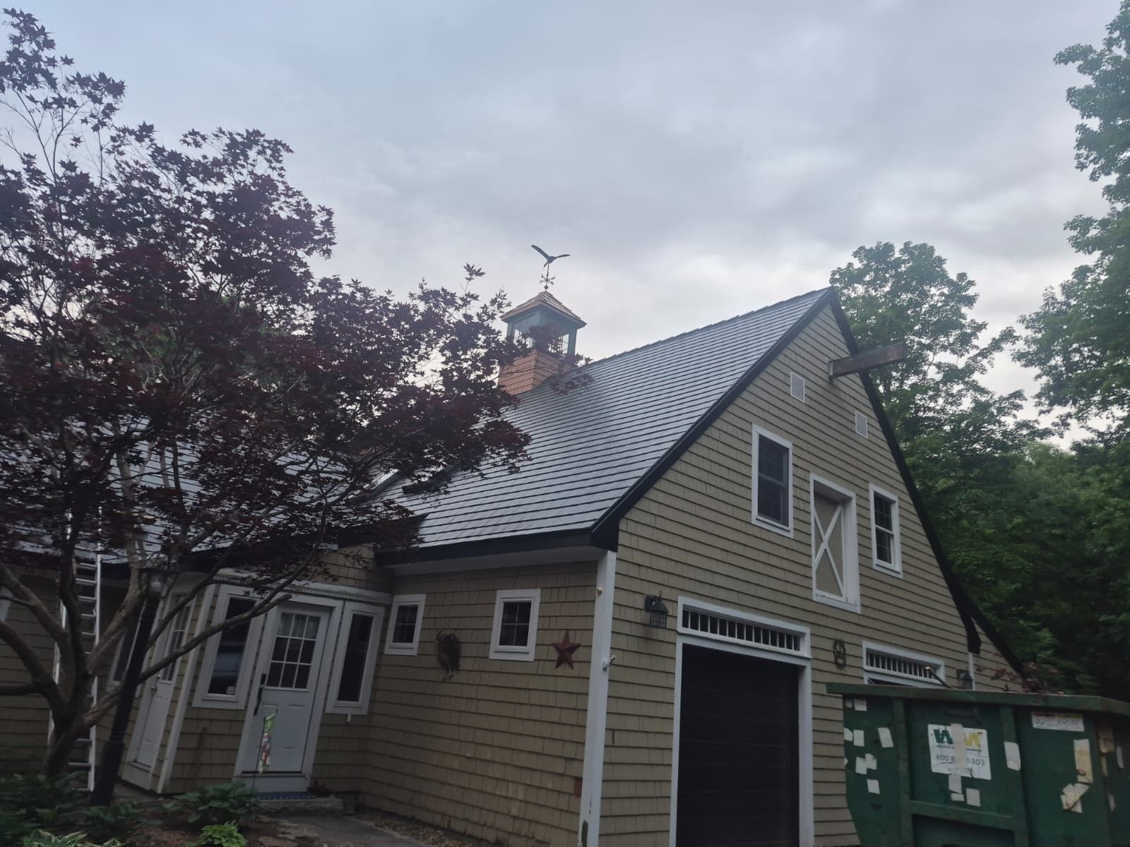 Tan tan house with steep metal roof, front porch, and garage beside trees at dusk