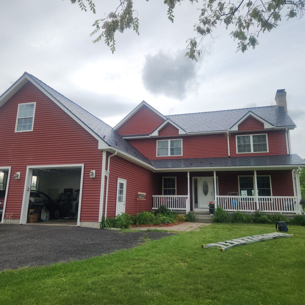 Red farmhouse with attached garage, white porch, and green lawn under a cloudy sky.