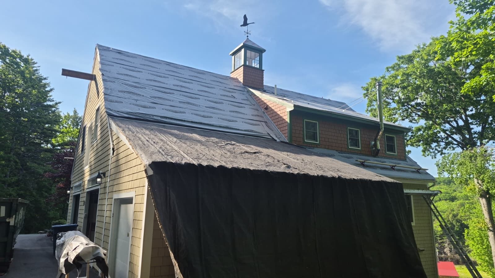 Stone building with a shingled roof and cupola, partly covered by a dark tarp under a blue sky.