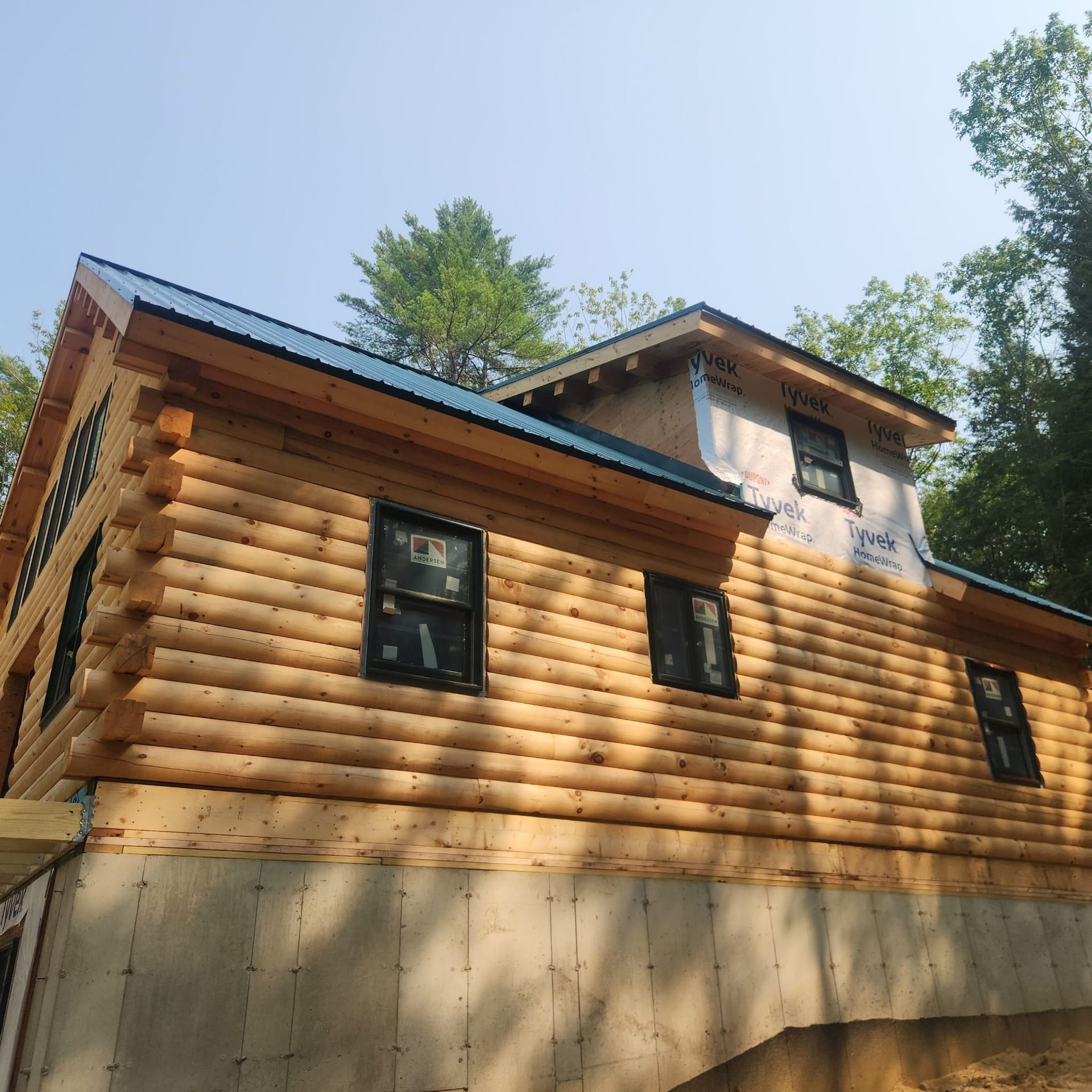 Wooden cabin with log siding, dark windows, and a concrete foundation surrounded by trees under a blue sky