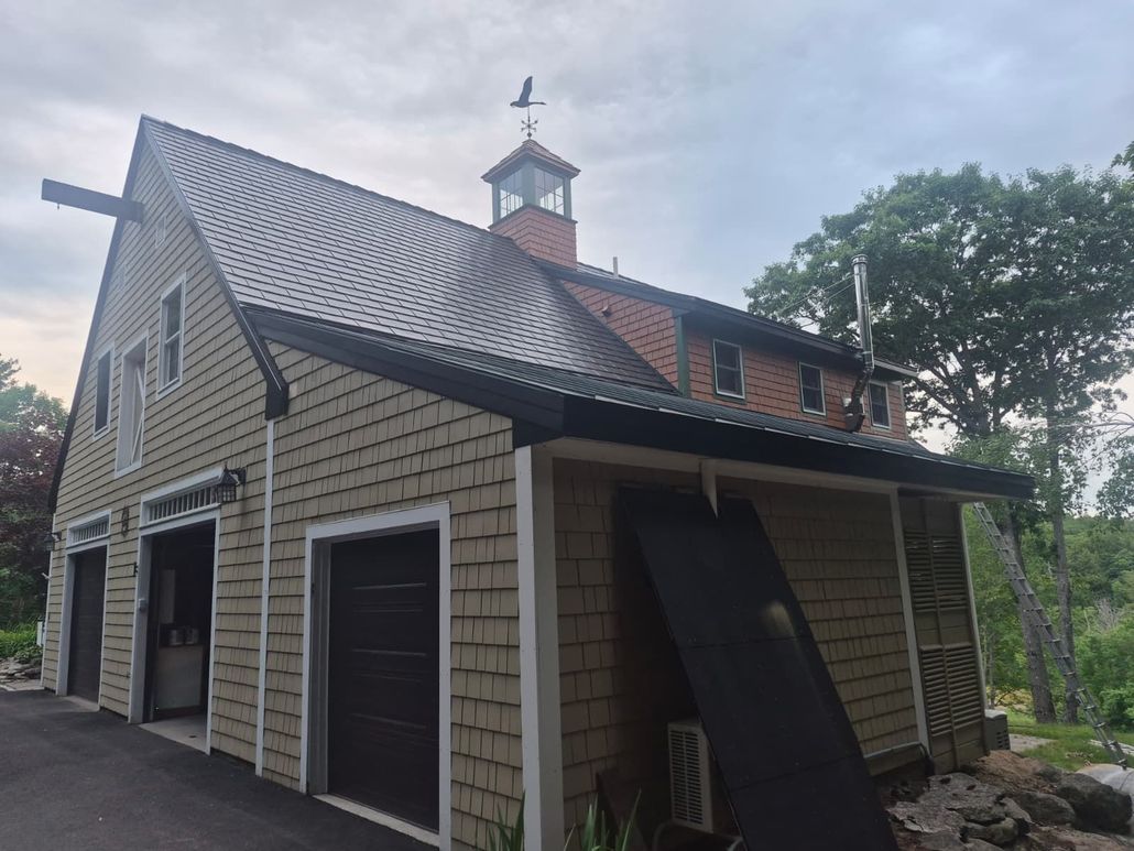 Gray house with steep metal roof, dormer windows, and attached garage in a wooded setting