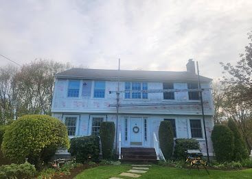 Large white two-story house with blue shutters, front porch, and trimmed hedges under a cloudy sky