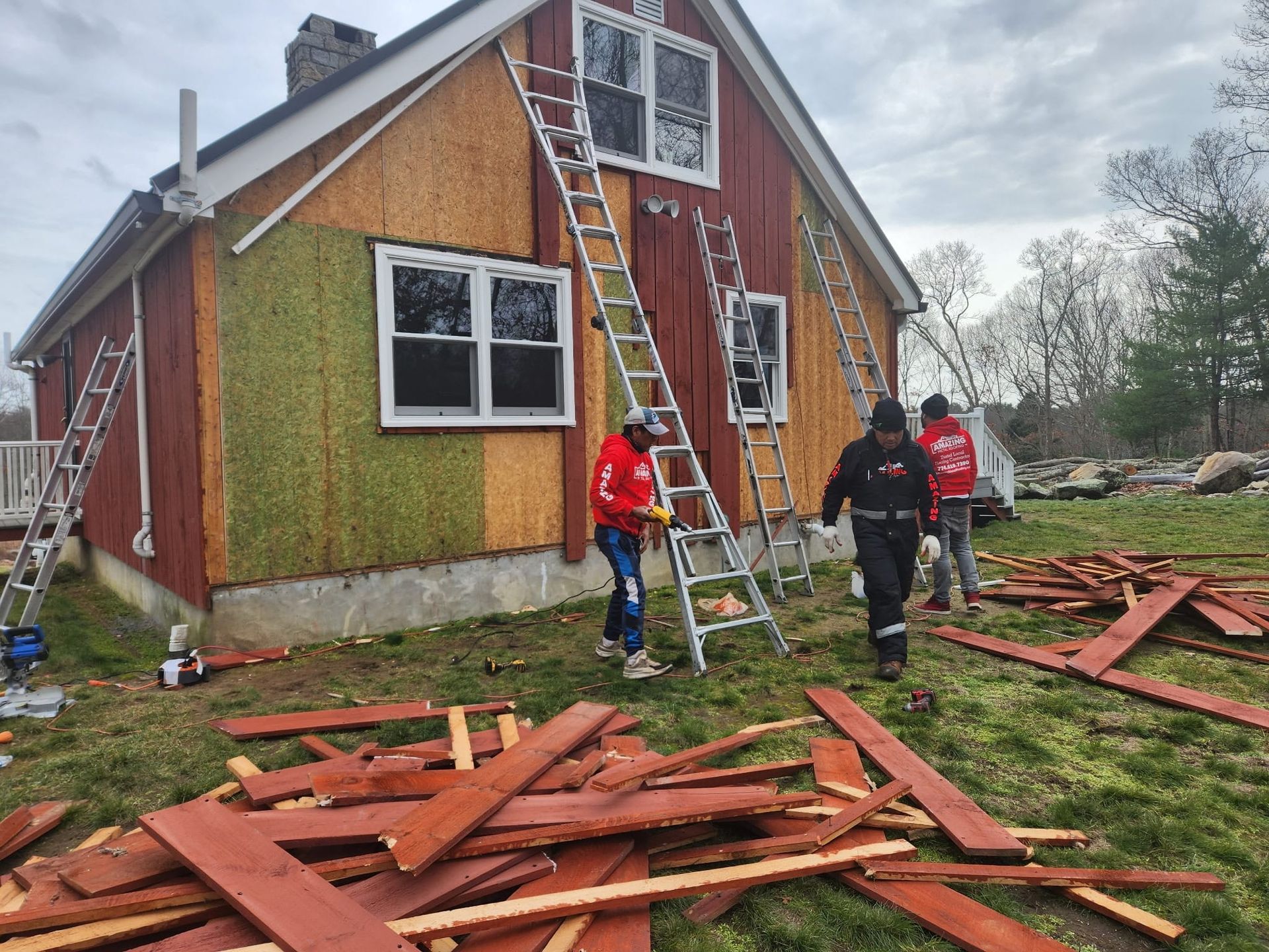 Workers on ladder repairing roof damage on a red house, with debris scattered on the lawn.