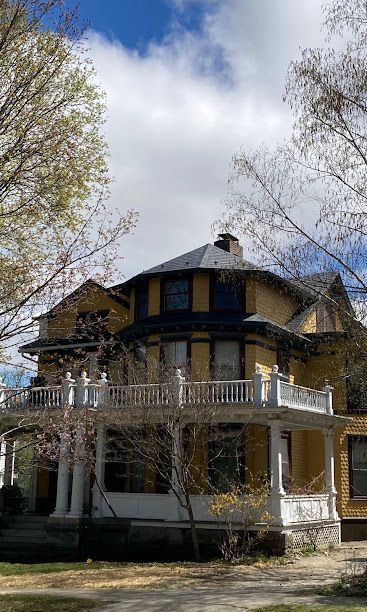 Victorian house with a wraparound porch and yellow trim under a partly cloudy sky