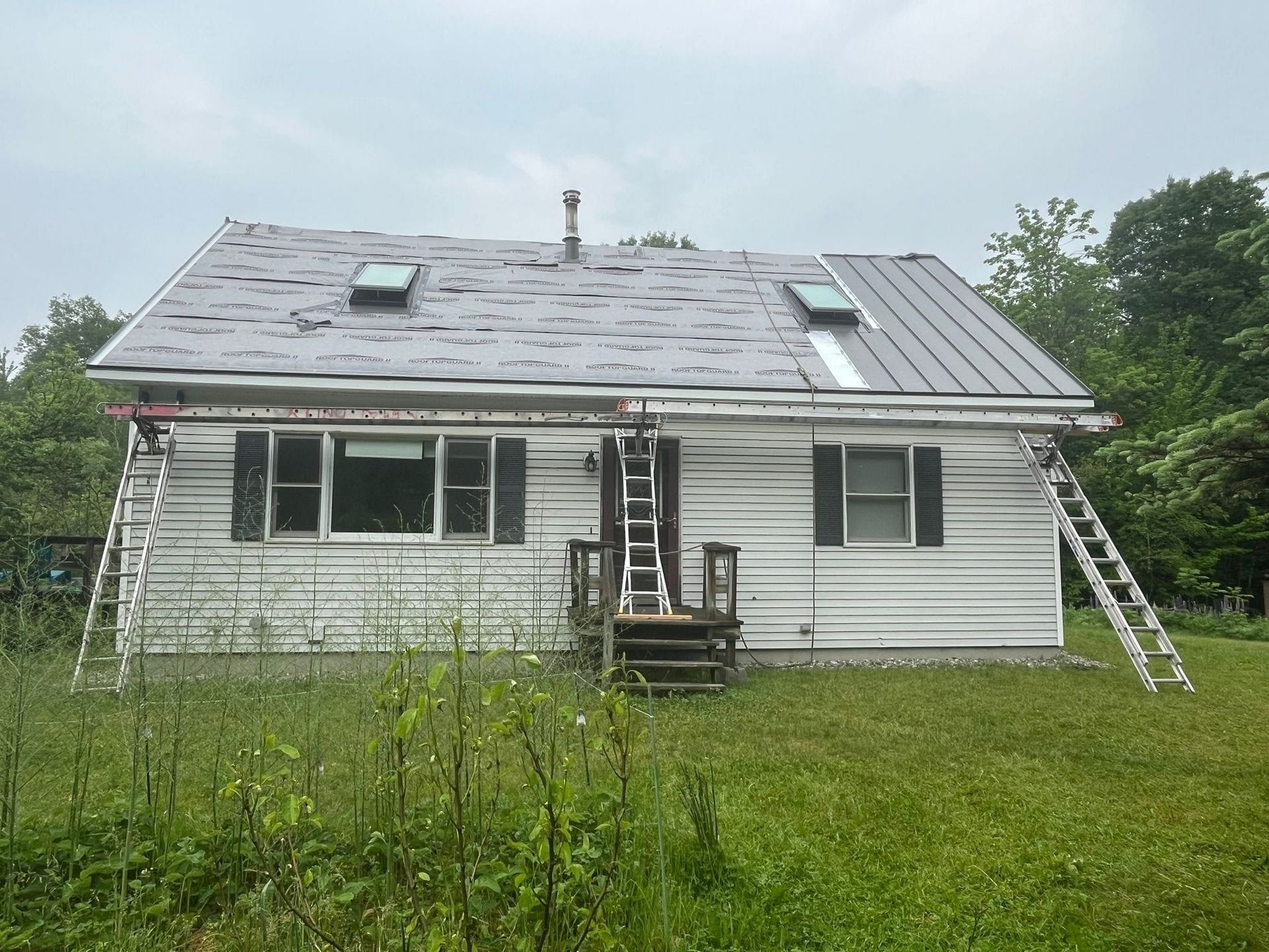 Small house with a gray metal roof, ladders on both sides, and overgrown grass in front