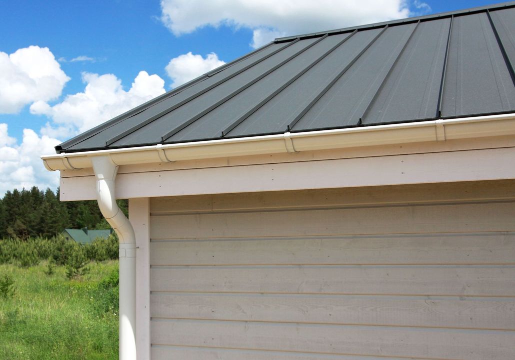Gray metal roof and white gutter on a beige building under a blue sky with clouds