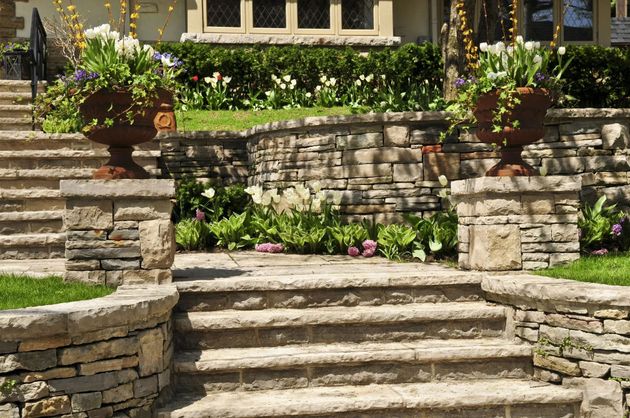Stone steps leading up to a house, flanked by stone walls and planters with flowers.