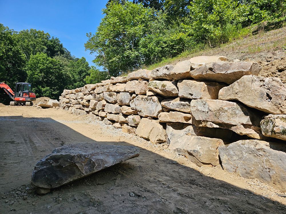 Stone retaining wall under construction next to a dirt road, with trees and an excavator in the background.