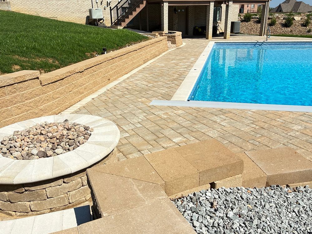 Poolside patio with fire pit, pavers, retaining wall with grass, and blue pool water.