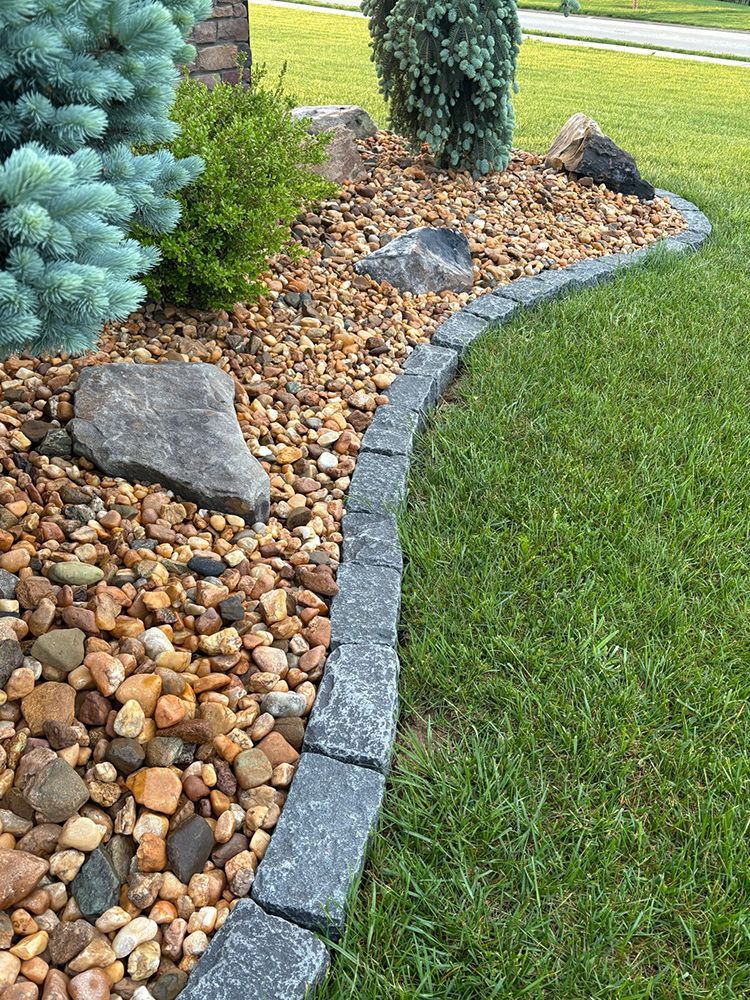 Landscape bed edged with dark stone, filled with gravel and plants, beside green grass.