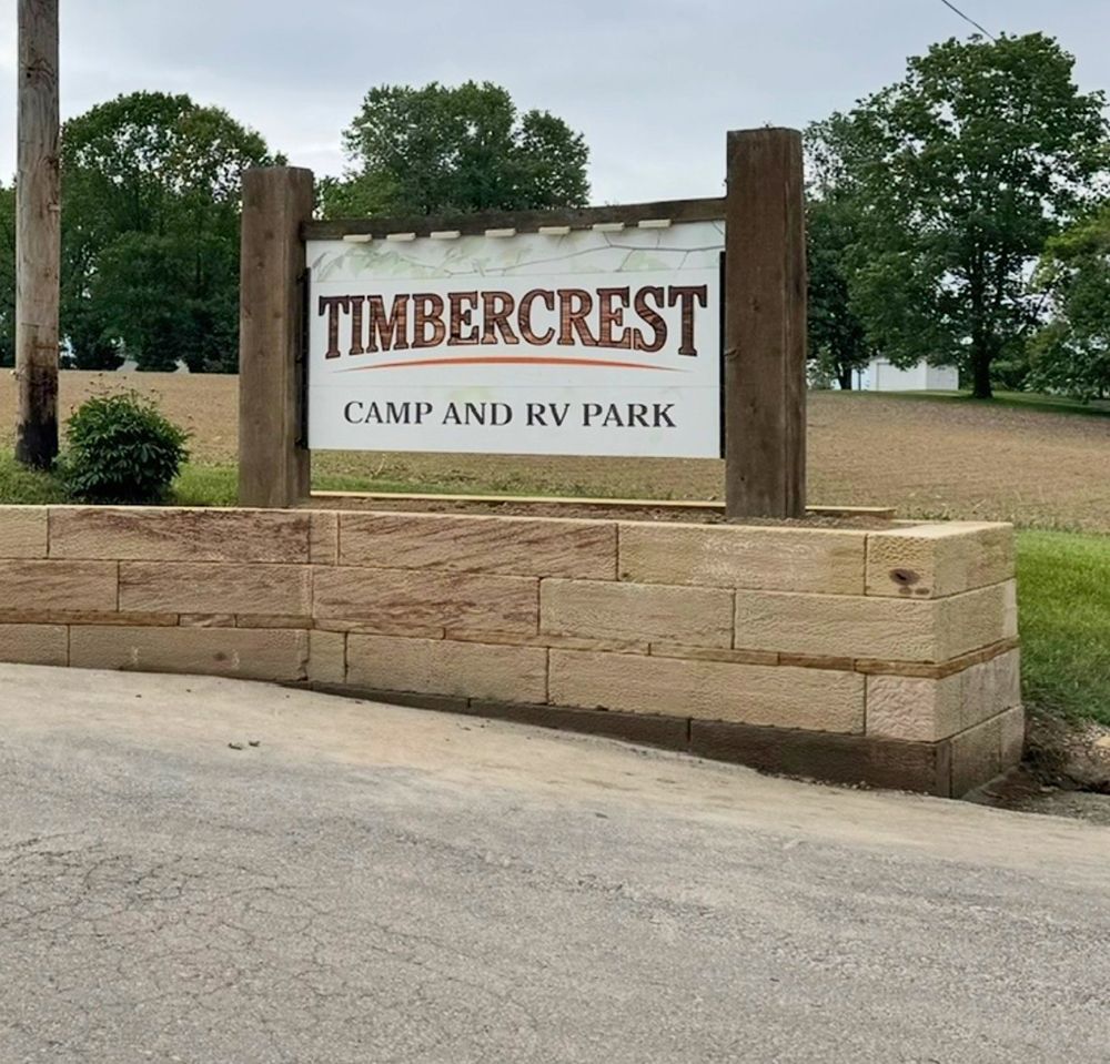Sign for Timbercrest Camp and RV Park. Brown sign on a brick wall with green trees in the background.
