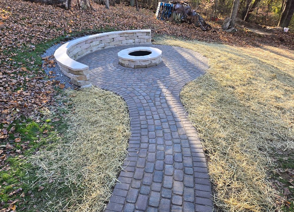 Brick path leading to a circular fire pit with a stone wall and surrounding dry grass.