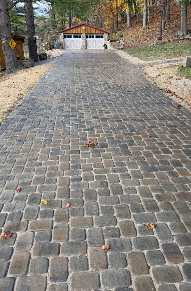 Cobblestone driveway leading to a two-car garage. Brown and gray pavers, leaves scattered.