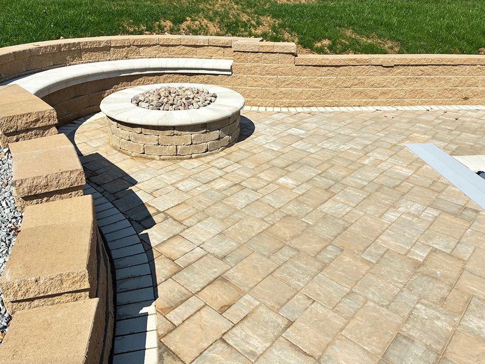 A brick patio with a circular fire pit and built-in stone seating, near a grassy area.