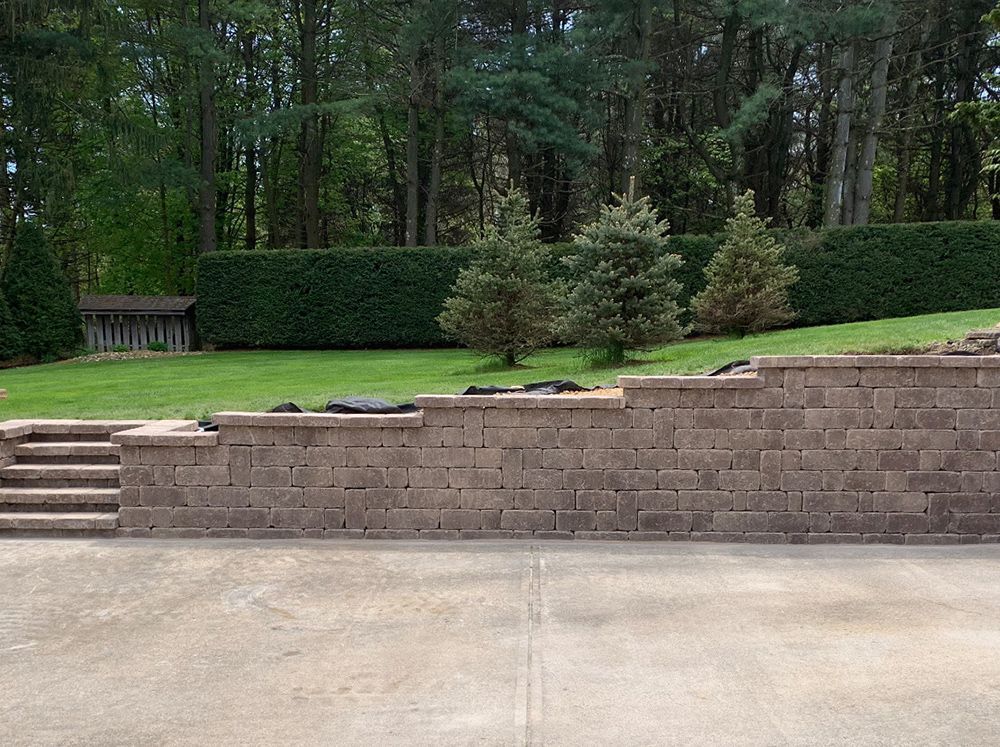 Retaining wall with steps leading to a grassy hill with evergreen trees against a backdrop of woods.