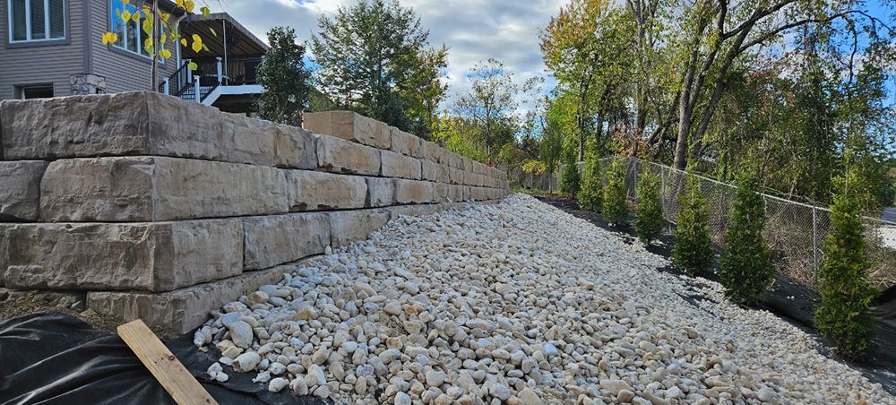 Stone retaining wall with gravel base, trees and building in the background.