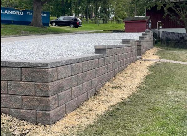 Gray retaining wall supports a gravel driveway; green grass in front.