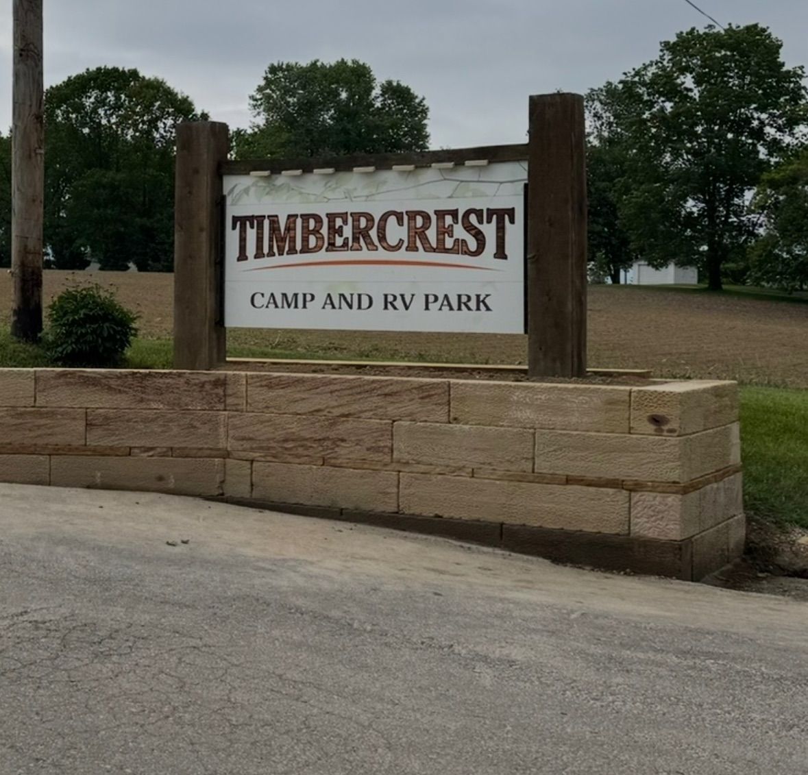 Sign for Timbercrest Camp and RV Park. Beige rectangular sign over a stone wall on a road, trees in the background.