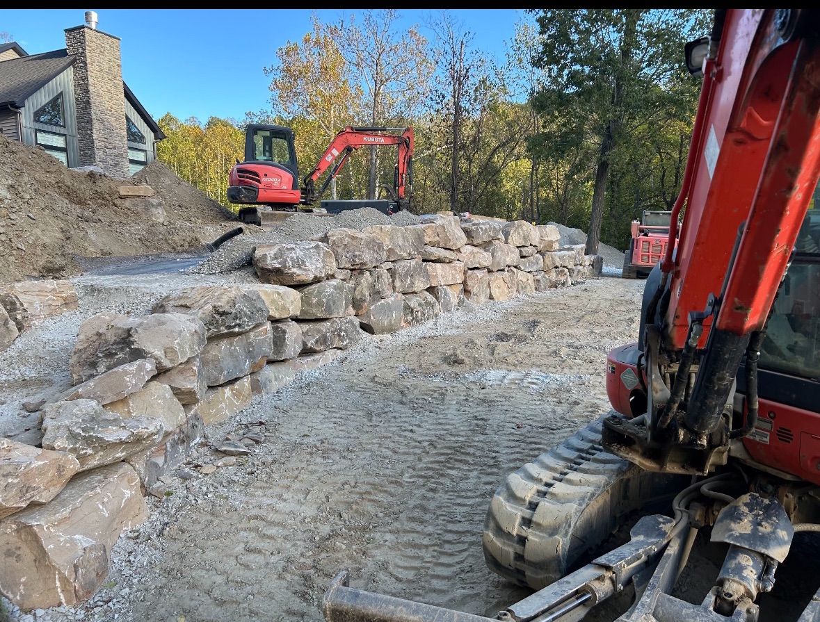 Construction site with two red excavators near a rock wall; dirt and trees in the background.