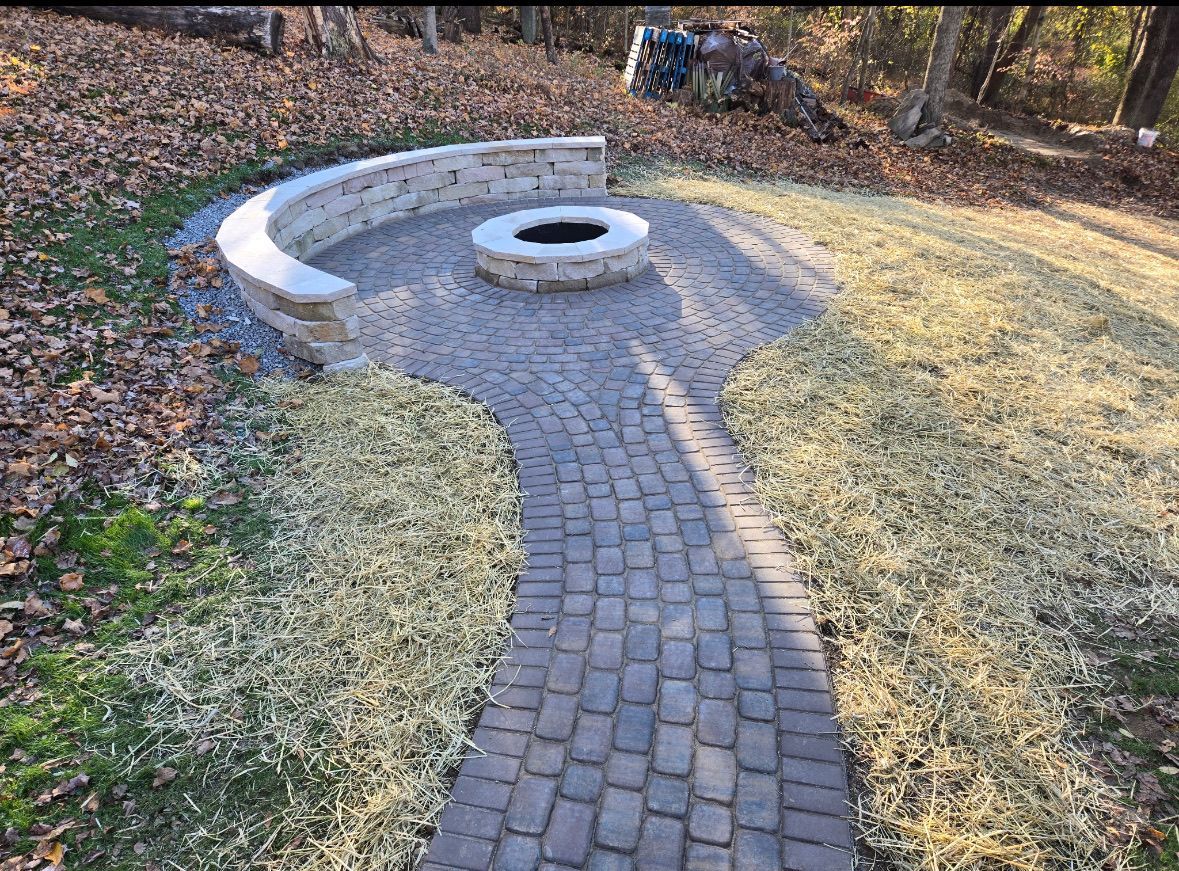 Brick pathway leading to a stone fire pit area with curved stone seating, surrounded by grass and leaves.