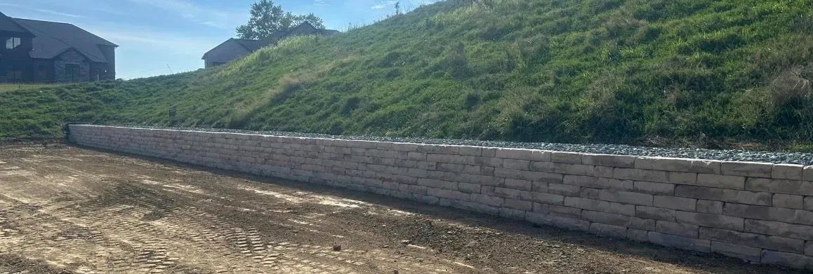 A retaining wall made of light-colored bricks sits between a grassy hill and an area of dirt and gravel.