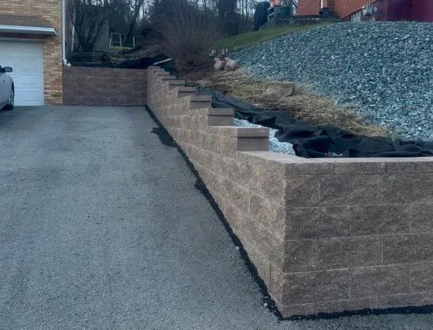 Stone retaining wall bordering a driveway, with a hillside of gravel and landscaping in the background.