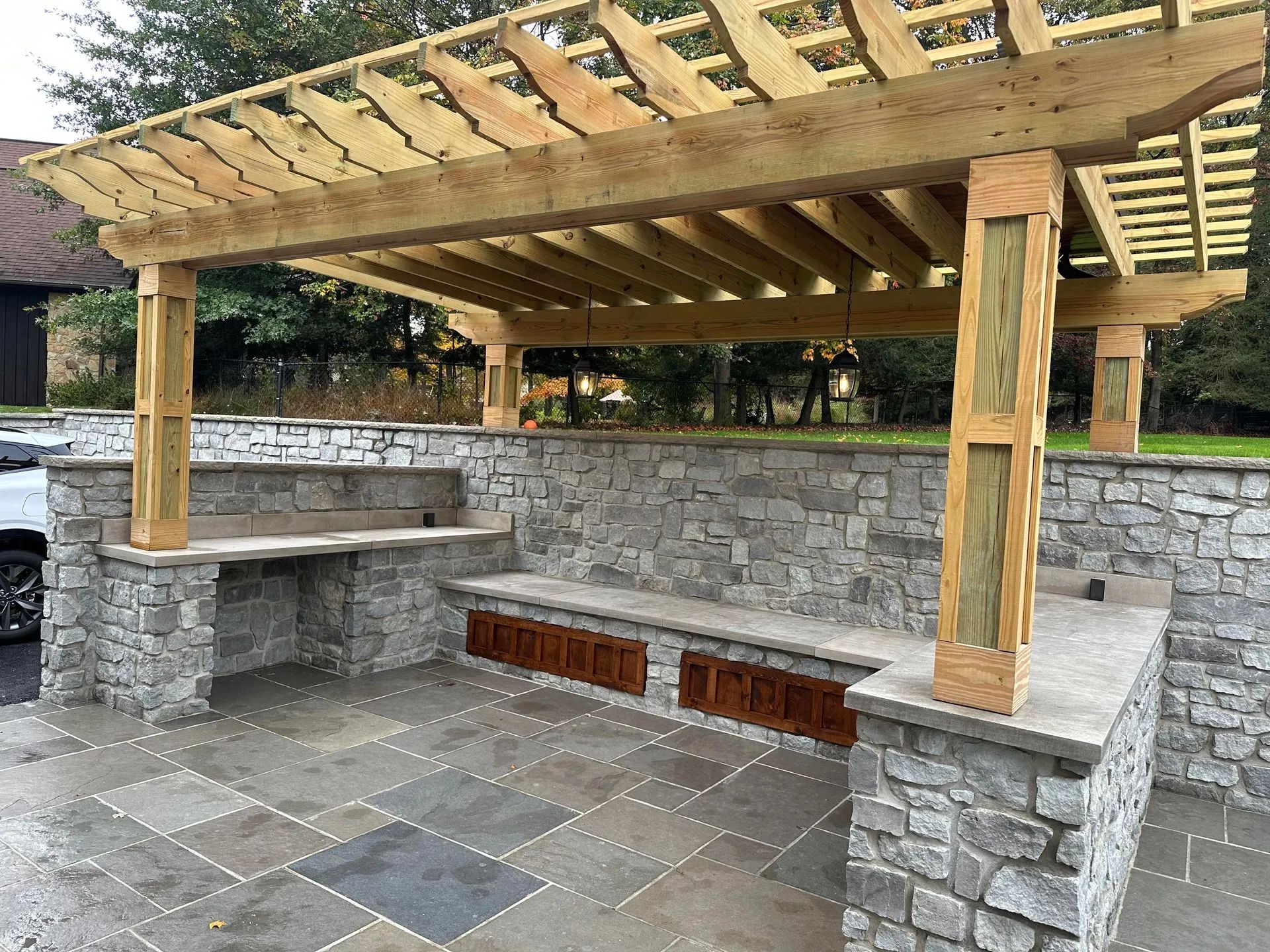 Stone-walled outdoor kitchen with pergola. Gray stone walls, wood pergola, and bench seating with brown cabinetry.