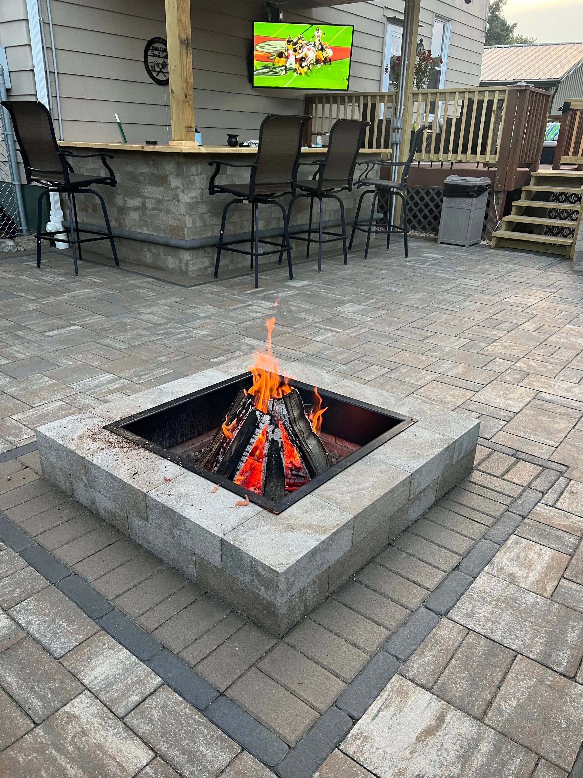 Outdoor fire pit with burning logs, surrounded by patio pavers. An outdoor bar and TV are in the background.