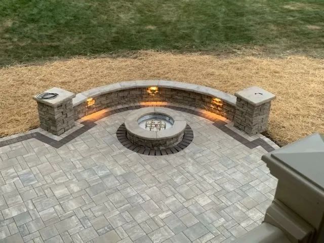 Stone patio with fire pit, curved wall with lights, and two pillars; surrounded by grass.