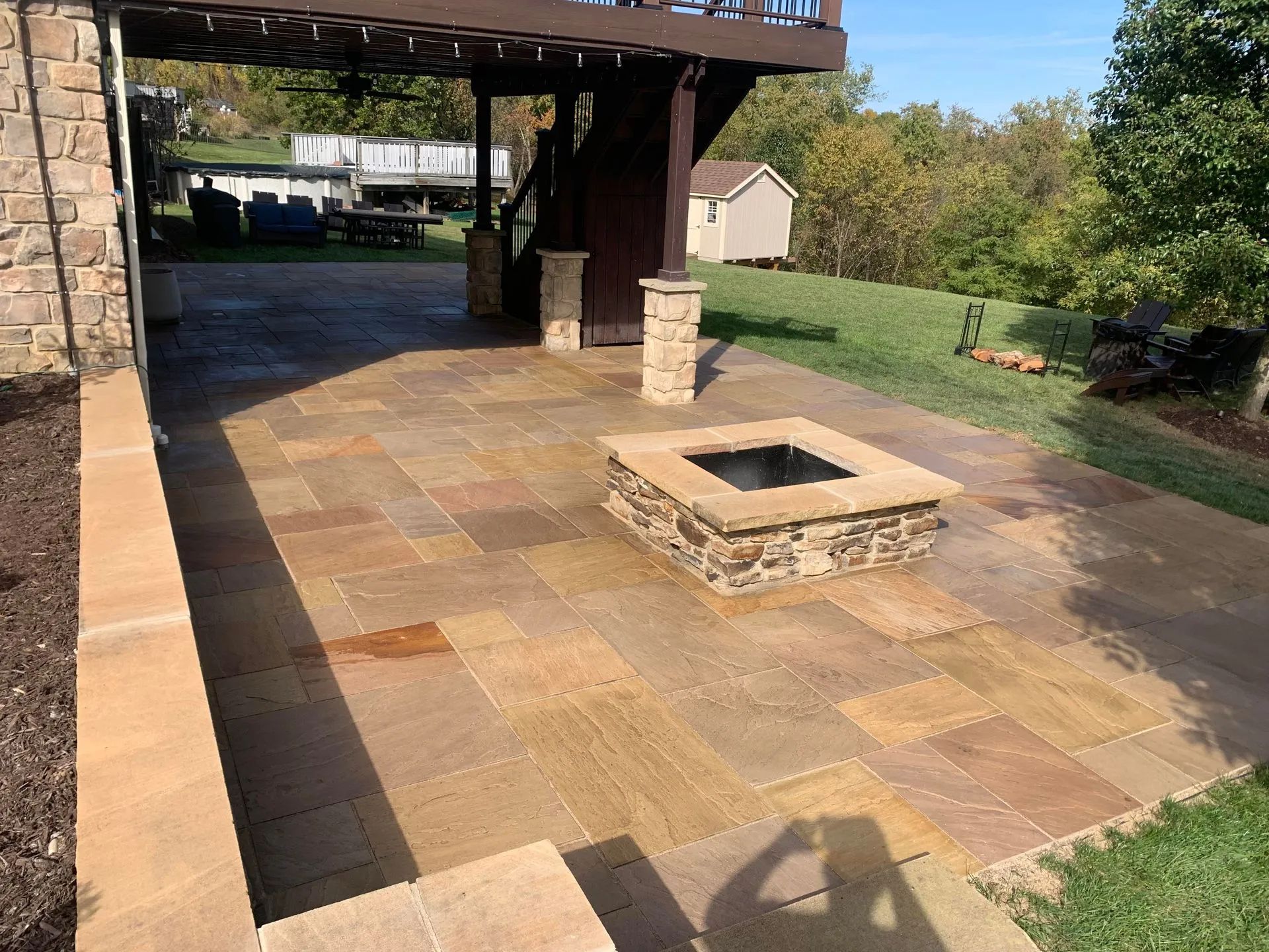 Stone patio with built-in fire pit, under a deck with green lawn and trees in the background.