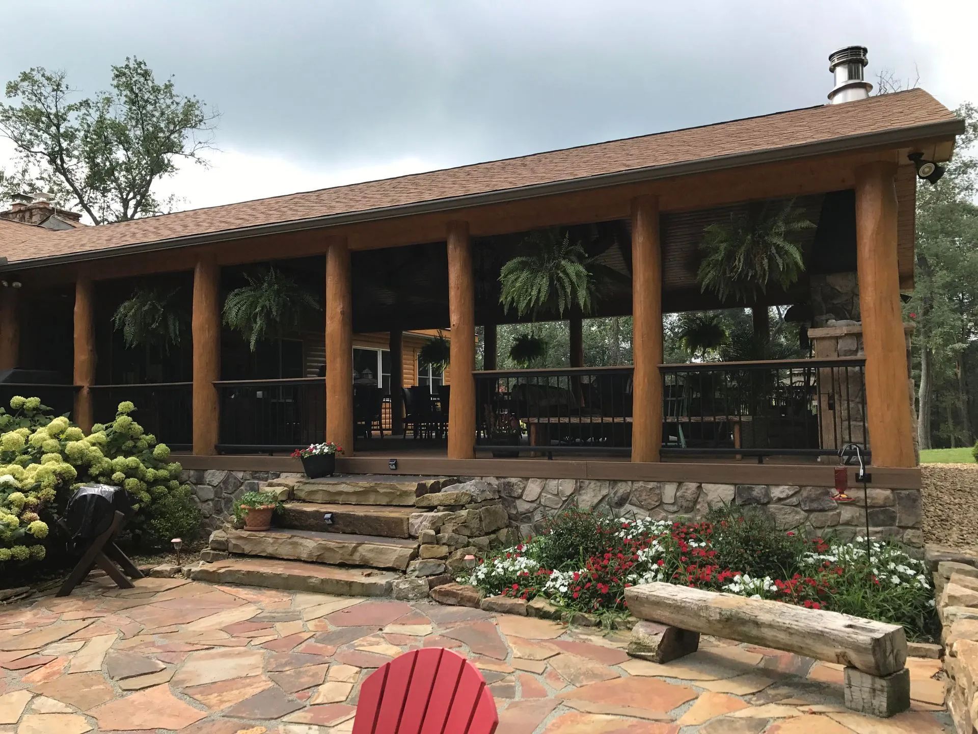 Screened-in porch with wood beams, stone base, and steps, overlooking a patio with a bench and plants.