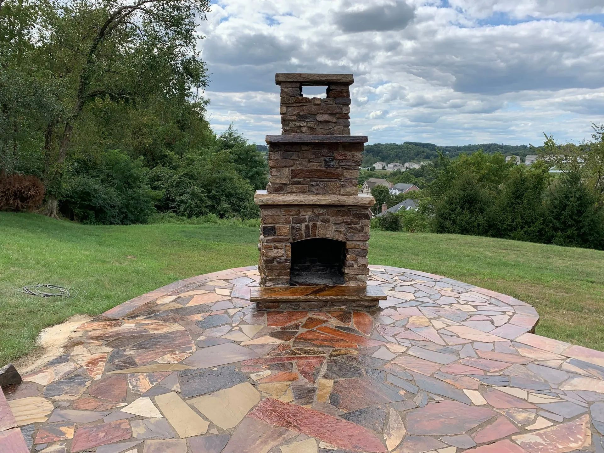 Stone outdoor fireplace on a colorful flagstone patio, with a grassy yard and a cloudy sky.