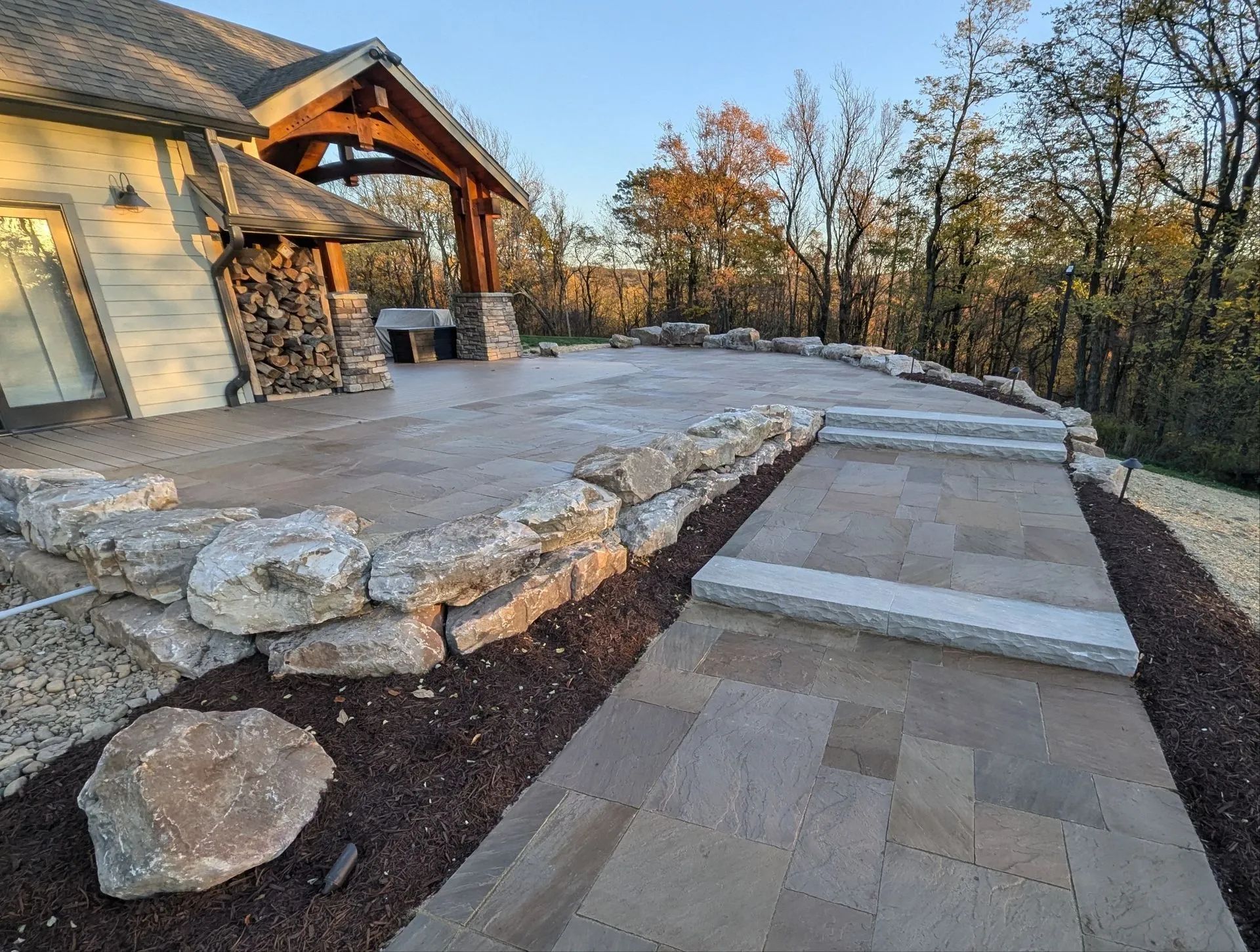 Stone patio with retaining wall, walkway, and steps leading to a house with wooden beams, surrounded by trees.