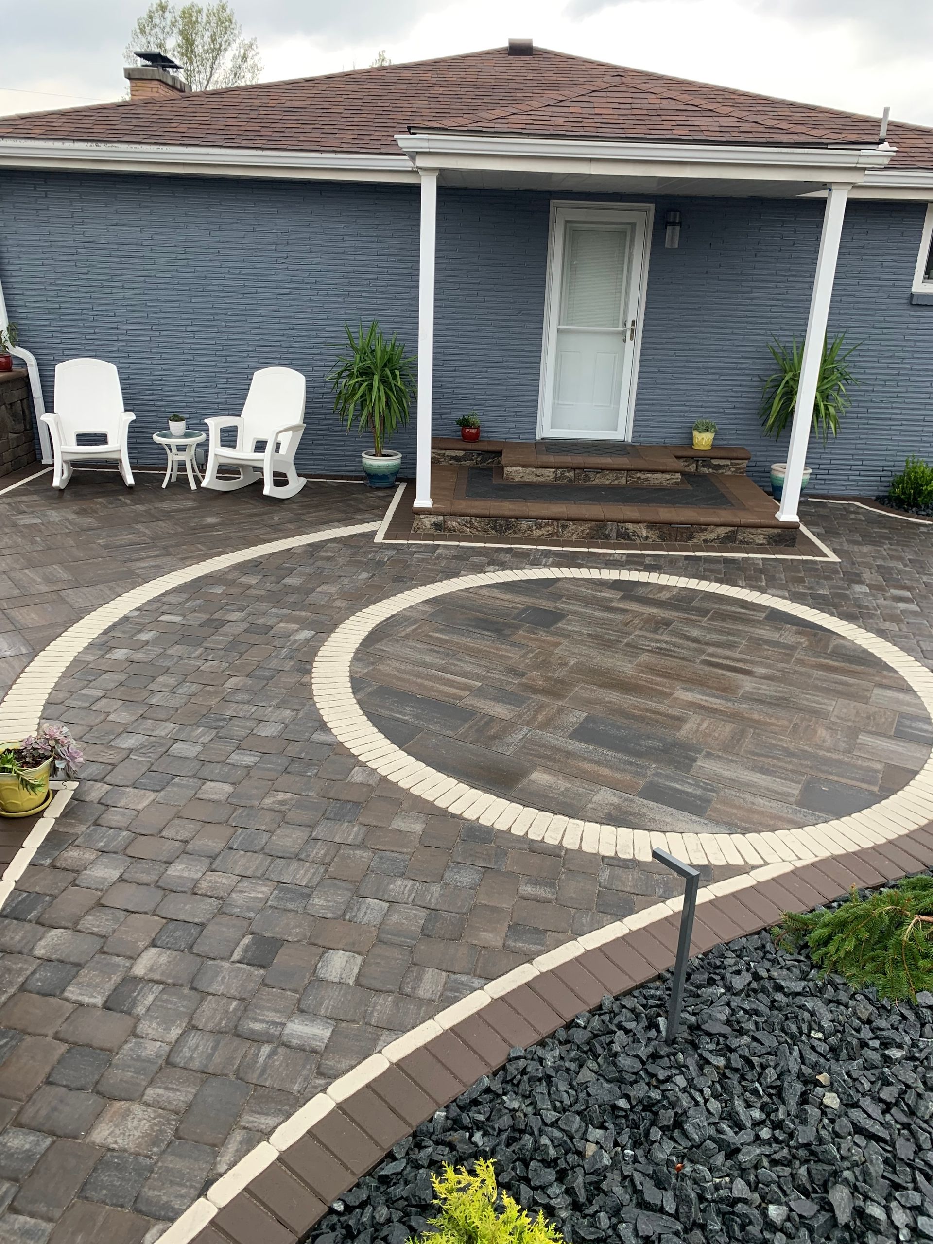 Brick patio with swirling design leading to a blue house with white chairs and a door.