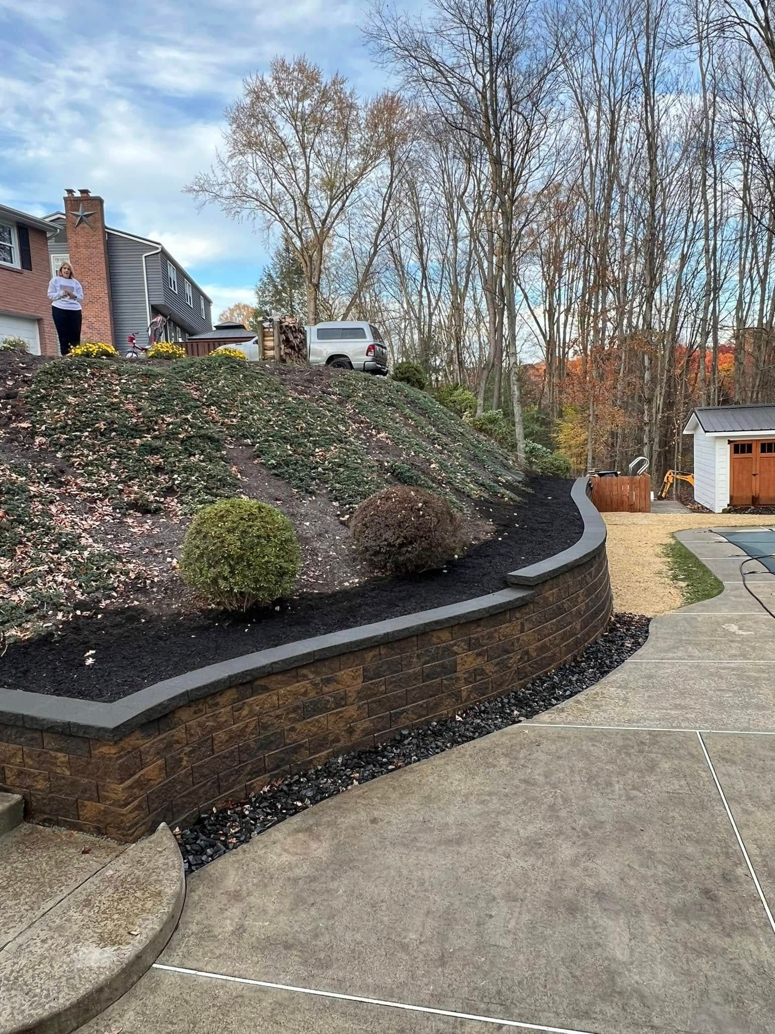 Concrete patio next to a retaining wall with landscaping. Person standing on hill in background.