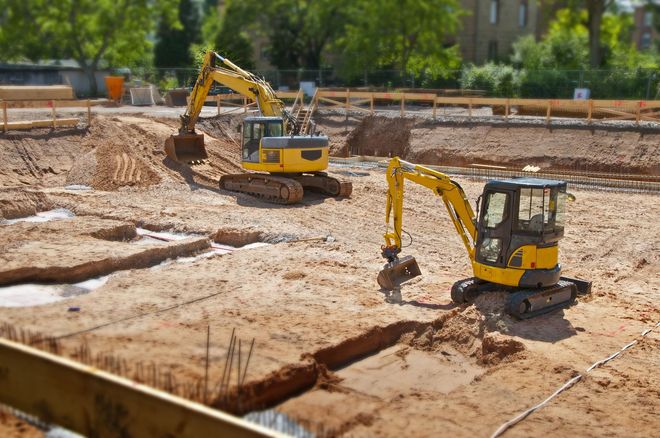 Two yellow excavators digging in a construction site; brown dirt, sunny day.