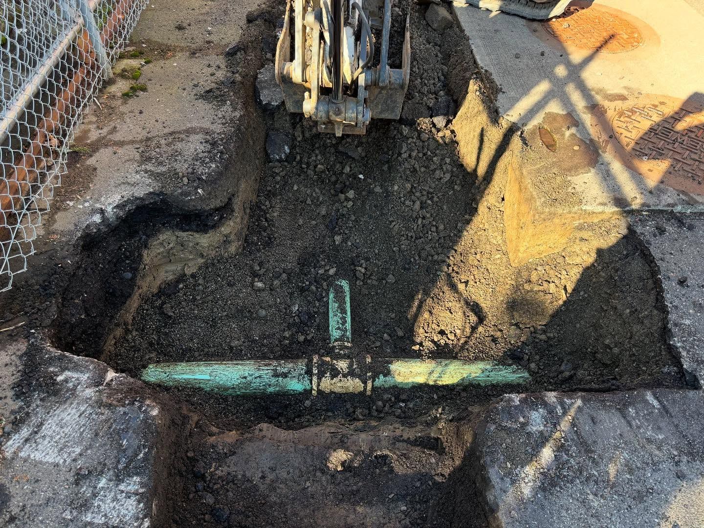 Trench dug around a copper pipe, with a small excavator above. Brown soil and asphalt surround.