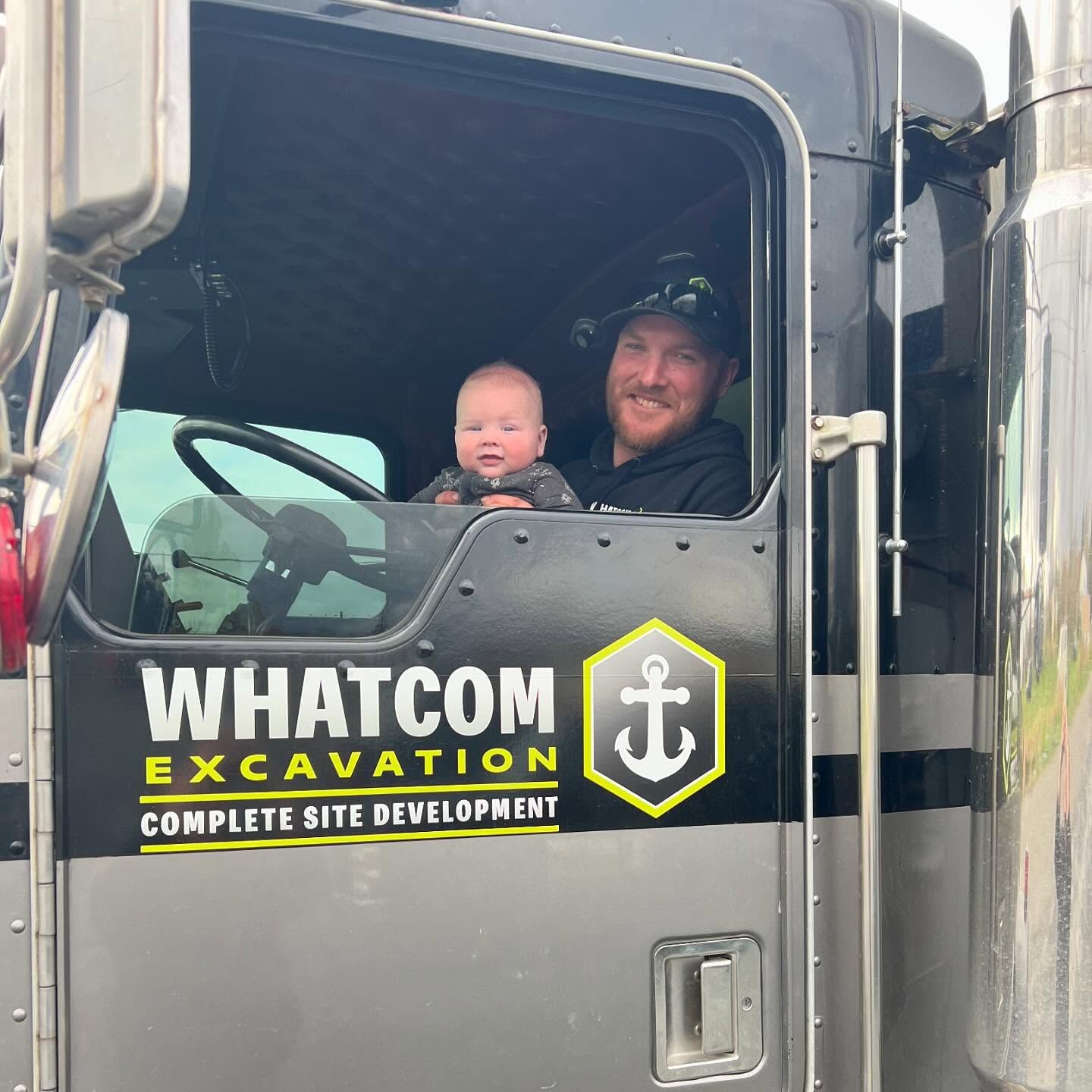 Man and baby in the cab of a Whatcom Excavation truck. Man smiles, baby looks at the camera.