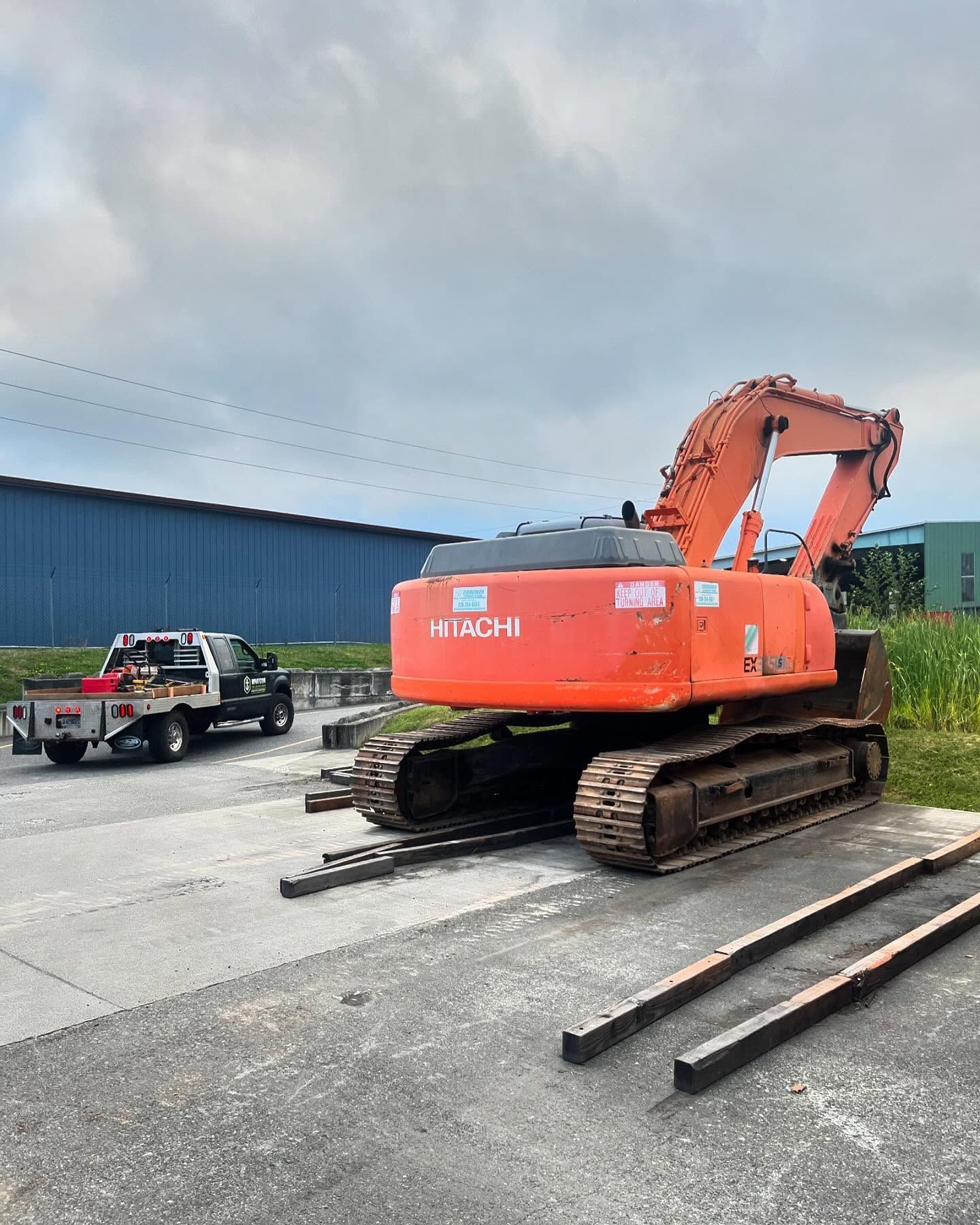 Orange Hitachi excavator parked next to a blue building and a pickup truck on a concrete surface.