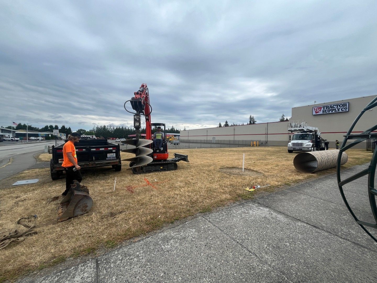 Construction site: a mini excavator drilling into the ground, worker and truck nearby, cloudy sky.
