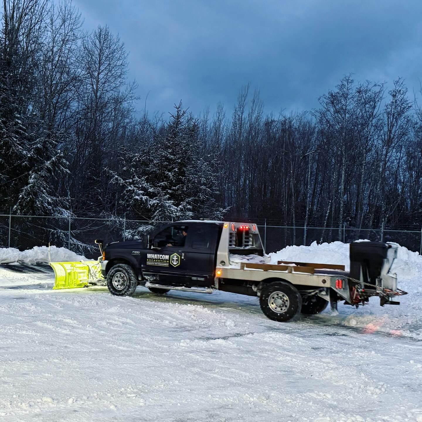Snowplow truck on a snow-covered surface with a yellow plow. Trees in the background, overcast sky.