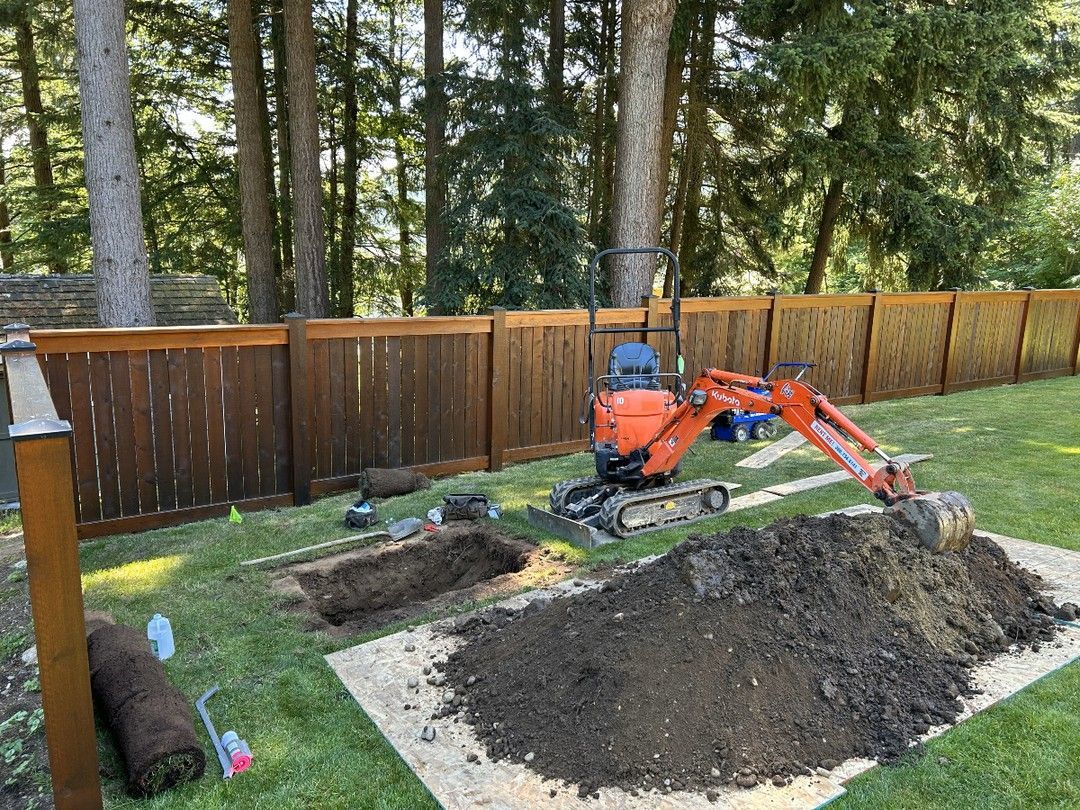 Mini excavator digging in a backyard with a wooden fence. Pile of dirt and dug-up area visible.