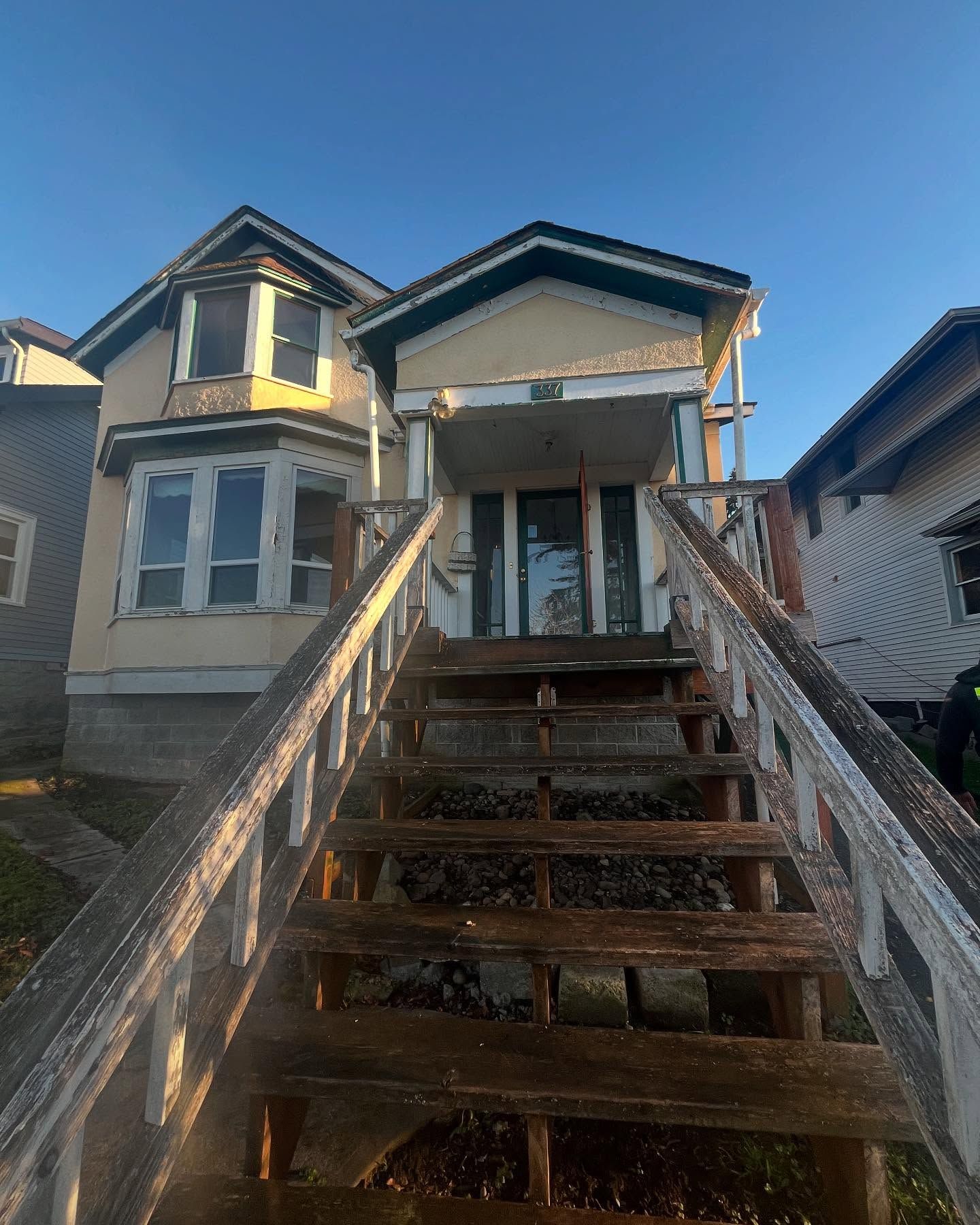 Dilapidated two-story house with wooden porch and stairs; weathered paint, overcast sky.