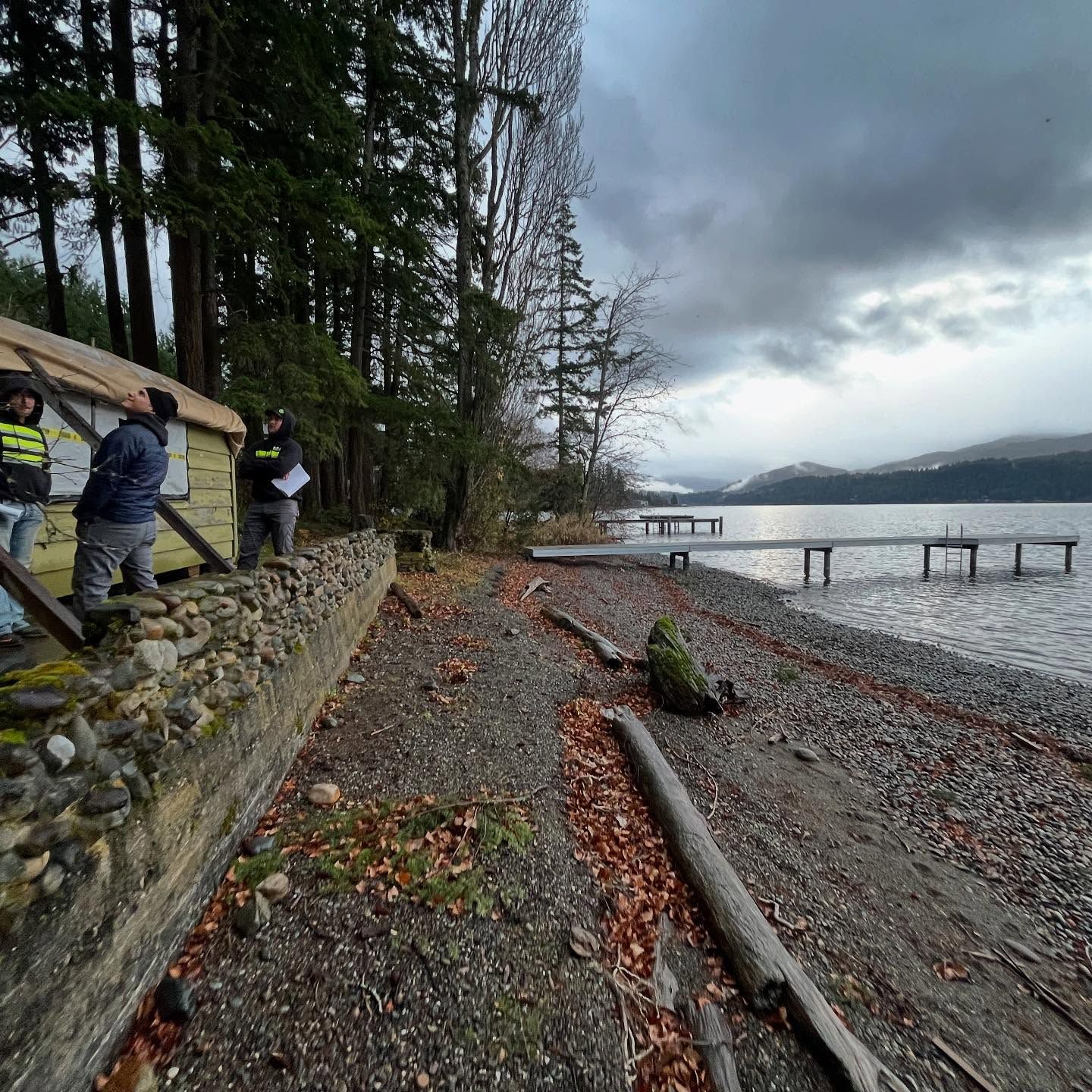 People on a shoreline, by a building and lake. Overcast skies.