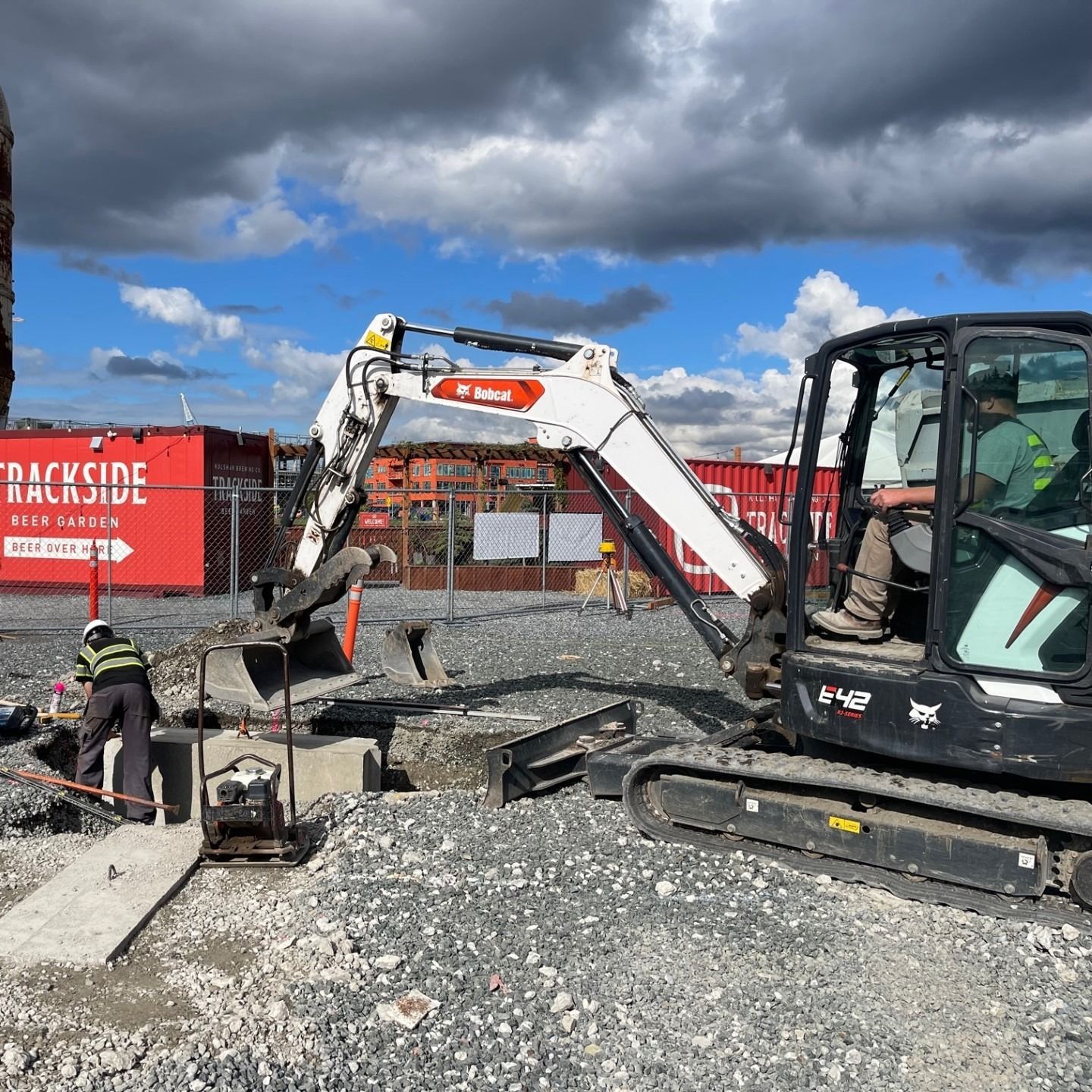 Construction site: Mini excavator and worker excavating concrete near red containers under cloudy sky.