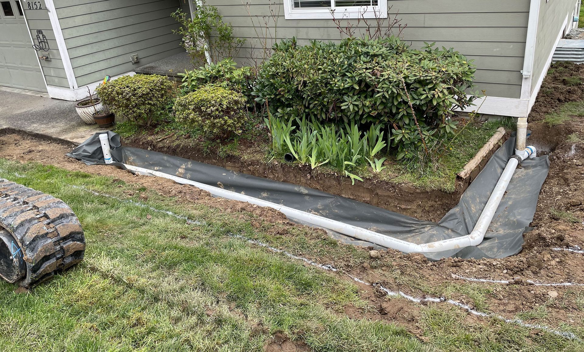 A trench lined with black fabric and white pipe for drainage near a house and garden.